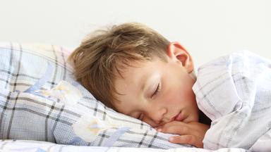 Auf dem Foto sieht man einen Jungen, der im Bett liegt und schläft. Seine Bettwäsche ist hellblau kariert, mit weißem Blumenmuster drauf.