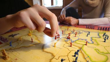 Auf dem Bild sieht man eine Hand und ein Spielbrett. Die Hand versetzt gerade eine Spielfigur.