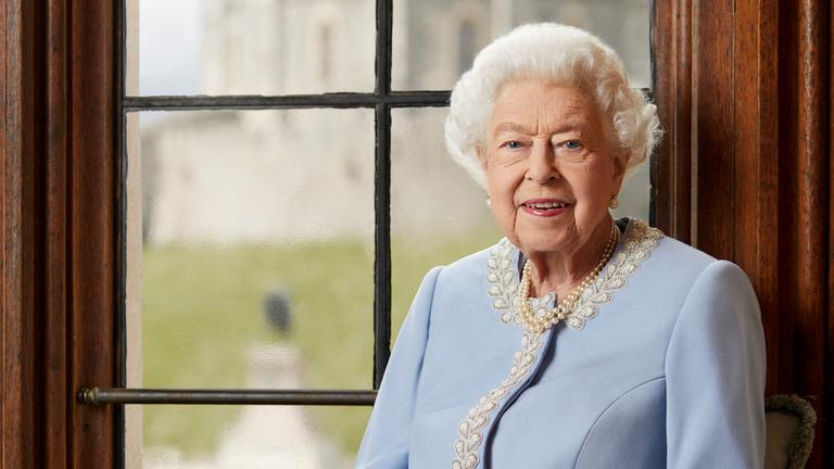 Rot uniformierte Sargträger tragen den blumenverzierten Sarg der verstorbenen Queen Elizabeth II vor schwarzem Hintergrund. Oben prangt eine Krone. 