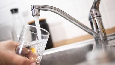 Ein Mann haelt ein Glas unter einen laufenden Wasserhahn.