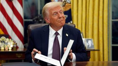 Donald Trump sitzt an seinem Schreibtisch im Oval Office, hinter ihm die Flagge der USA. Er klappt gerade einen Ordner zu und grinst zur Seite.