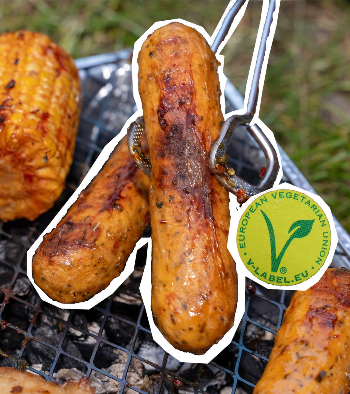 Auf dem Bild sind fleischlose Bratwürste auf einem Grill zu sehen. Eine wird von einer Grillzange gehalten. Im Hintergrund liegt noch ein Maiskolben sichtbar. Das Symbol für vegetarische und vegane Produkte ist neben die Würste eingefügt.