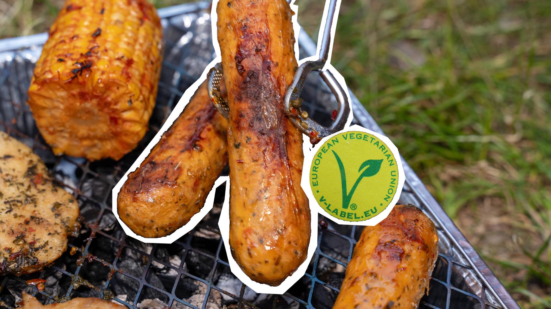Auf dem Bild sind fleischlose Bratwürste auf einem Grill zu sehen. Eine wird von einer Grillzange gehalten. Im Hintergrund liegt noch ein Maiskolben sichtbar. Das Symbol für vegetarische und vegane Produkte ist neben die Würste eingefügt.