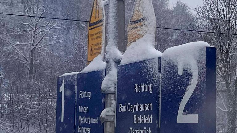 Nahaufnahme verschneiter Gleisbeschilderung mit den Nummern 1 und 2. Richtungsangaben nach Minden, Nienburg Weser, Hannover sowie Bad Oeynhausen, Bielefeld, Osnabrück. Schilder, Mast und Halterungen stark mit Schnee bedeckt.