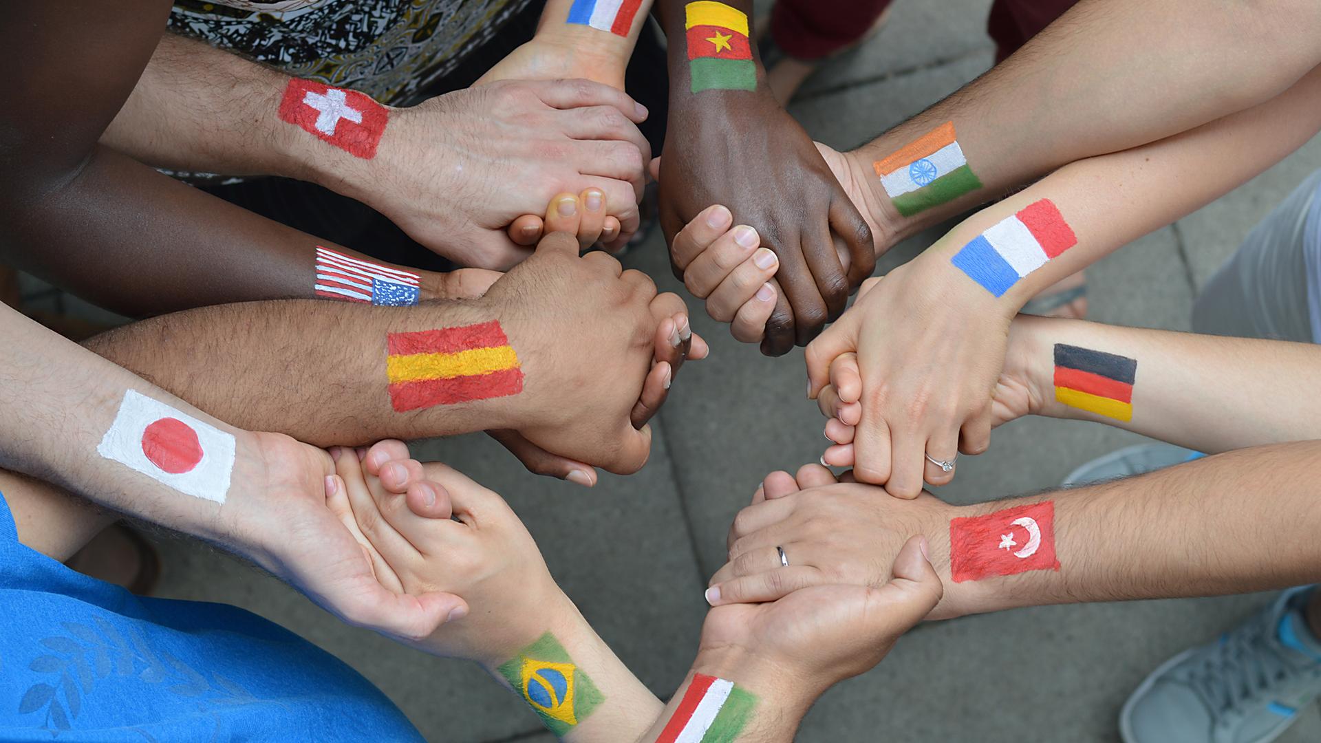 Viele Hände halten sich gegenseitig. Auf die Handgelenke sind Flaggen unterschiedlicher Länder gemalt.