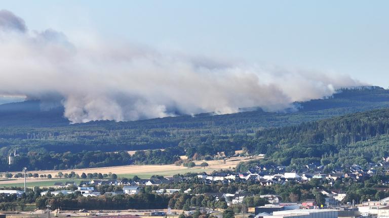 Panoramaaufnahme: Landschaft mit Häusern, aus dem Wald im Hintergrund steigen große Rauchwolken.