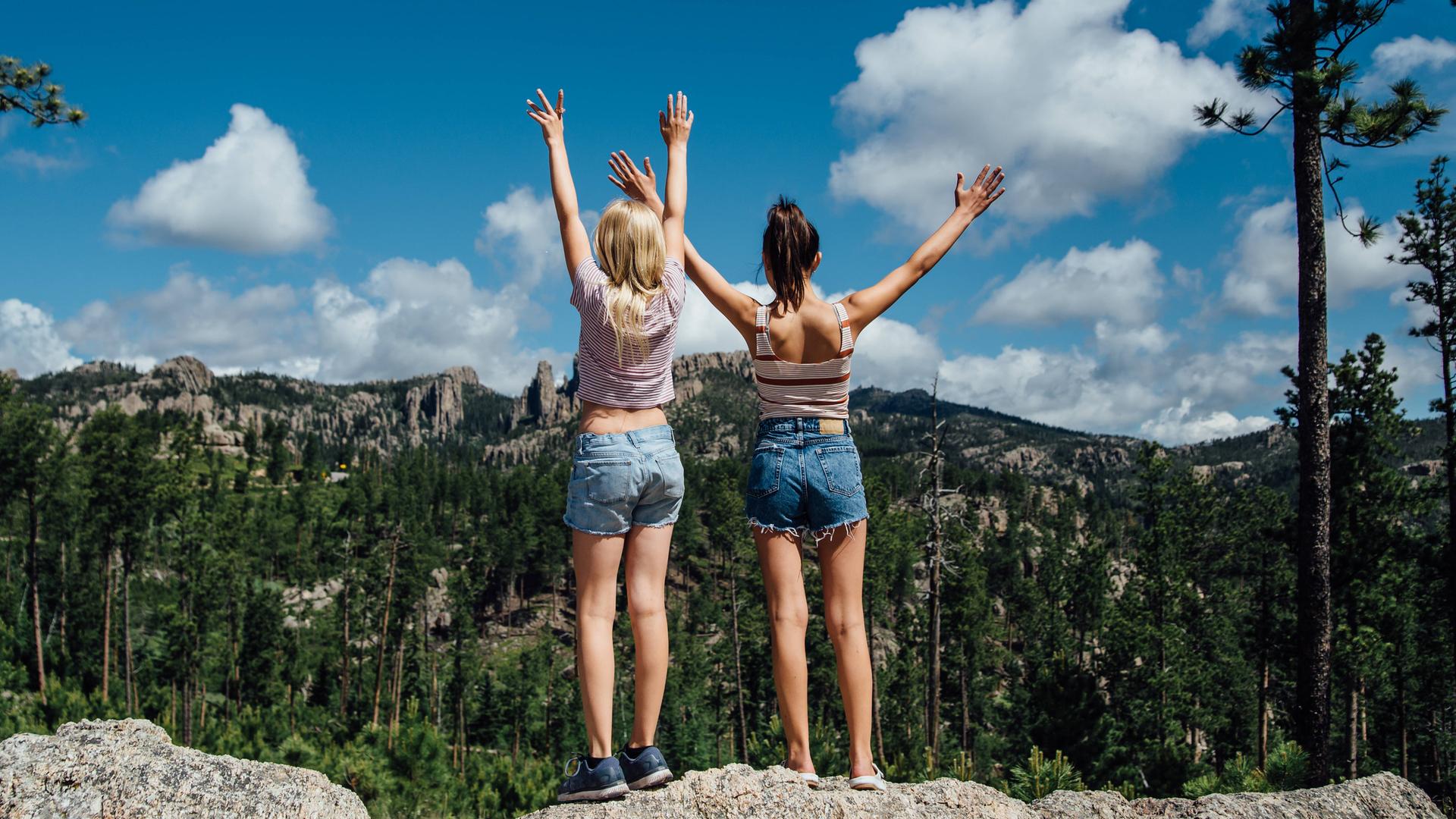 Zwei Mädchen stehen auf einem Felsvorsprung und strecken die Hände in den Himmel.