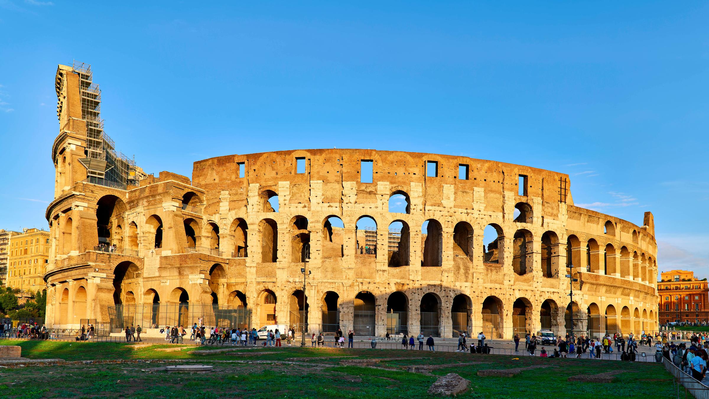 Das Kolloseum vor blauem Himel. Vor dem Kolloseu laufen Menschen.