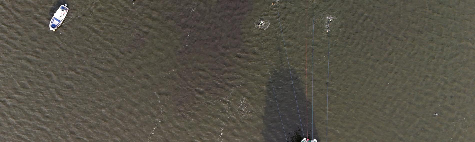 „Von oben sieht man einen See mit drei Booten und fünf Schwimmern. Die Schwimmer ziehen ein großes grünes Schiff durchs Wasser.“