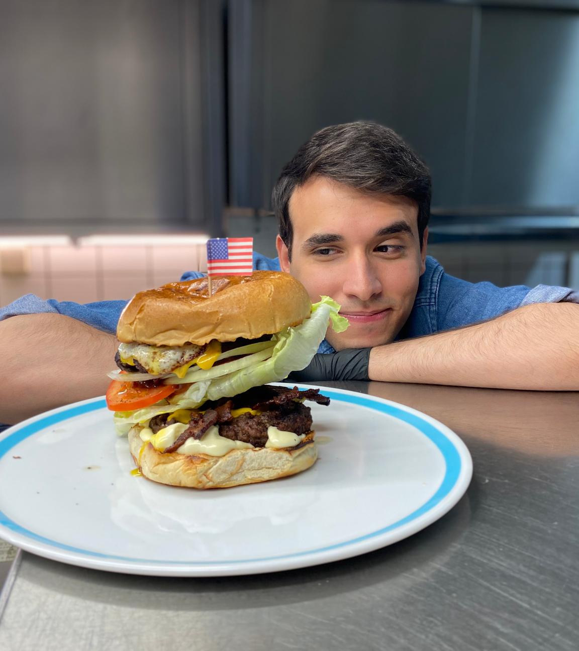 Auf dem Foto sieht man Logo-Reporter Sherif. Er stützt seinen rechten Arm und seinen Kopf auf der metallenen Arbeitsfläche in einer Großküche ab. Gleichzeitig schaut er auf den Teller vor sich, auf dem ein großer Hamburger liegt. Oben drauf auf dem Burger steckt eine kleine Flagge der USA aus Papier. Sherif guckt dem Hamburger stolz an.