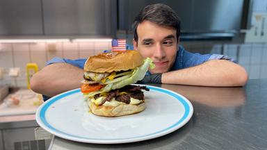Auf dem Foto sieht man Logo-Reporter Sherif. Er stützt seinen rechten Arm und seinen Kopf auf der metallenen Arbeitsfläche in einer Großküche ab. Gleichzeitig schaut er auf den Teller vor sich, auf dem ein großer Hamburger liegt. Oben drauf auf dem Burger steckt eine kleine Flagge der USA aus Papier. Sherif guckt dem Hamburger stolz an.
