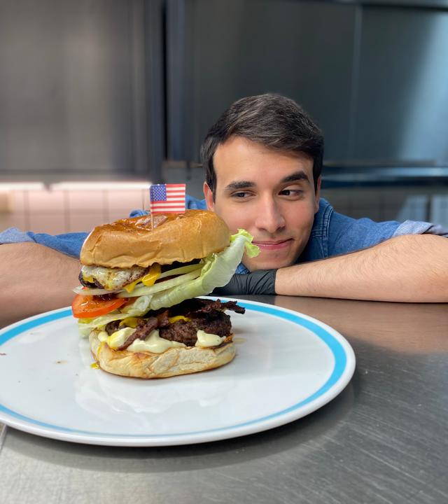 Auf dem Foto sieht man Logo-Reporter Sherif. Er stützt seinen rechten Arm und seinen Kopf auf der metallenen Arbeitsfläche in einer Großküche ab. Gleichzeitig schaut er auf den Teller vor sich, auf dem ein großer Hamburger liegt. Oben drauf auf dem Burger steckt eine kleine Flagge der USA aus Papier. Sherif guckt dem Hamburger stolz an.