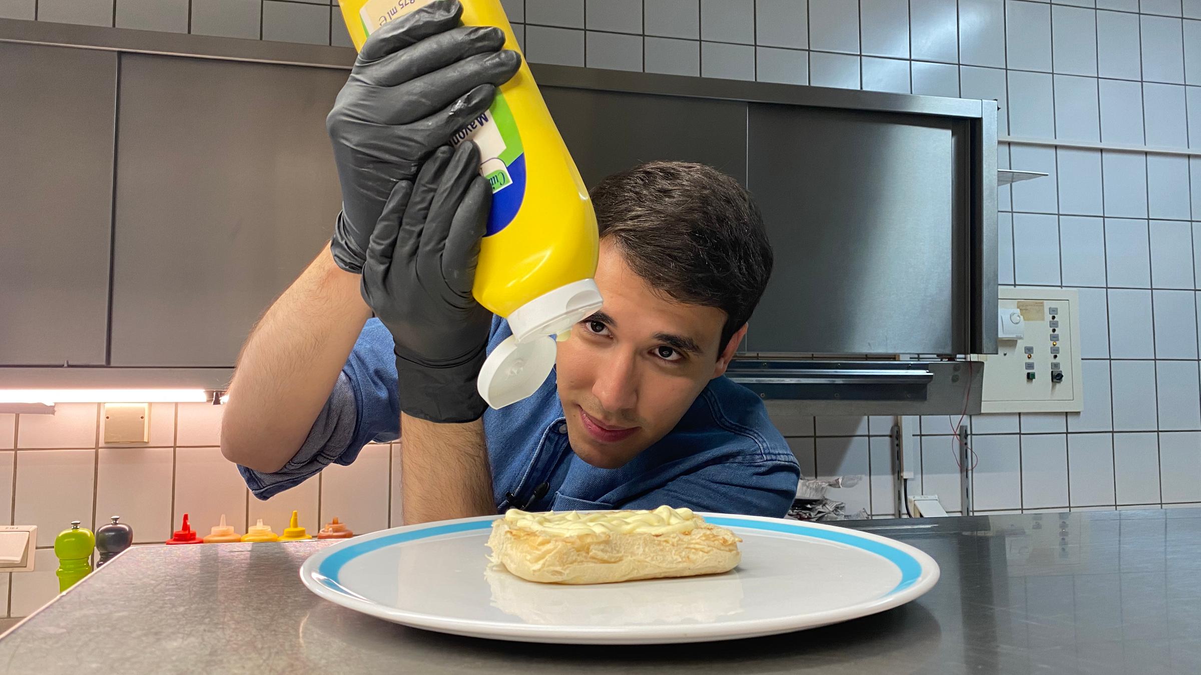 Auf dem Foto sieht man Sherif, wie er eine große Mayonnaise-Flasche über die untere Hälfte eines Burgerbrötchens hält. Dabei schaut er sehr konzentriert.