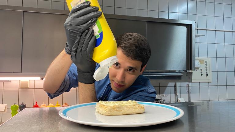 Auf dem Foto sieht man Sherif, wie er eine große Mayonnaise-Flasche über die untere Hälfte eines Burgerbrötchens hält. Dabei schaut er sehr konzentriert.