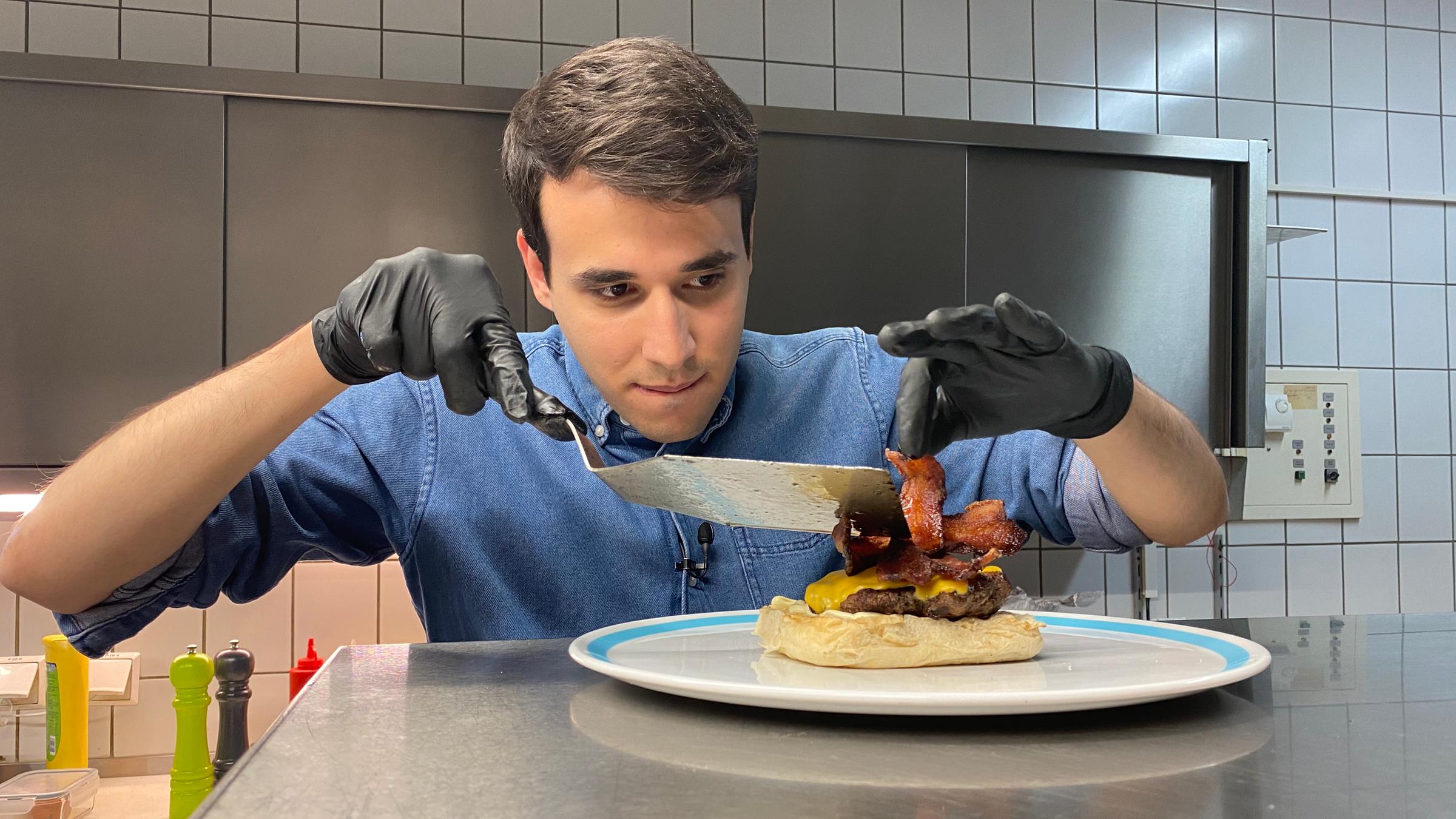 Auf dem Foto sieht man Sherif, wie er sehr konzentriert 2 Speck-Streifen auf den Hamburger legt. Vorher hat er schon ein Burgerpatty mit Käse überbacken und es auch auf die untere Hälfte des Burgerbrötchens gelegt. 