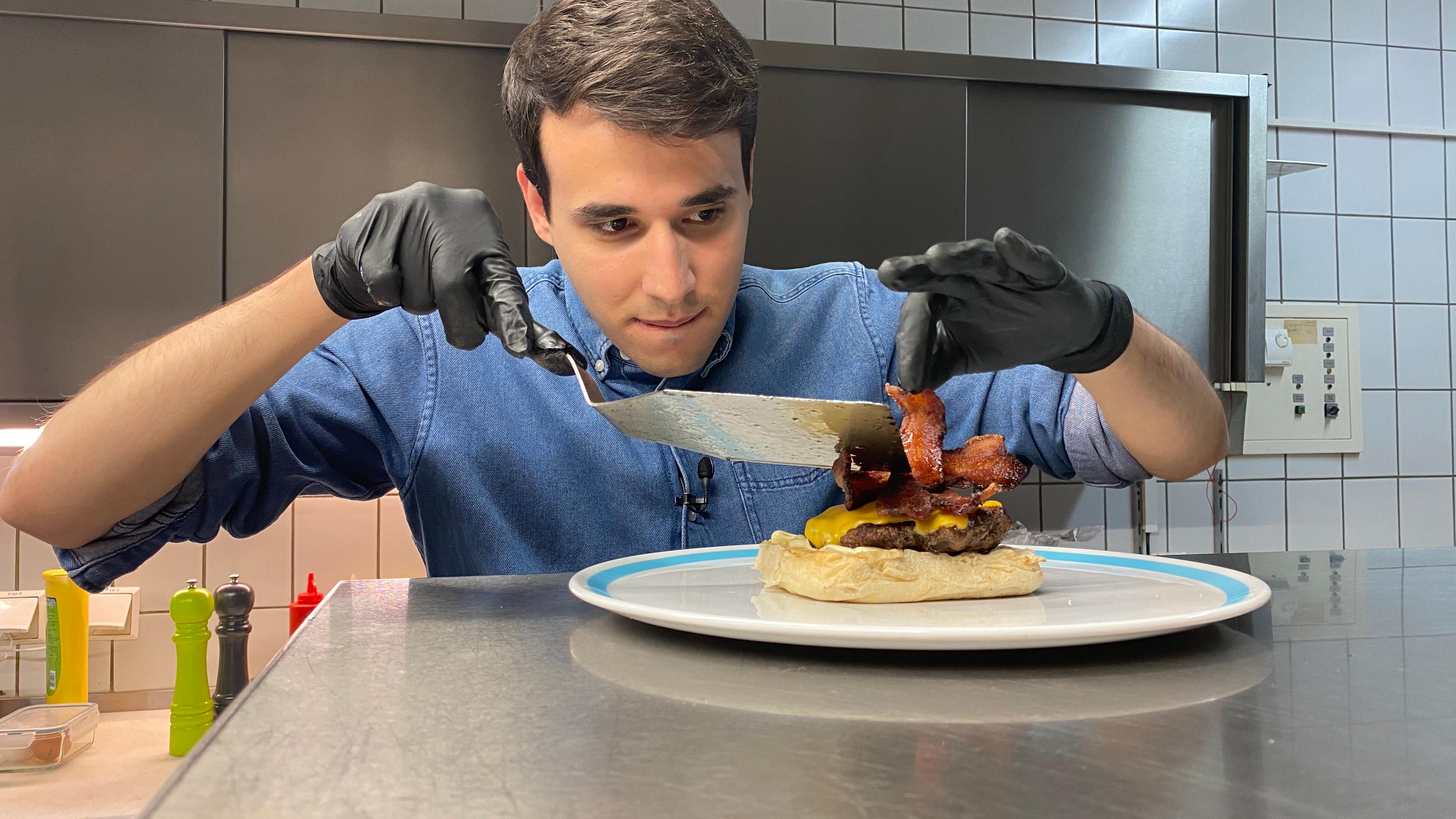 Auf dem Foto sieht man Sherif, wie er sehr konzentriert 2 Speck-Streifen auf den Hamburger legt. Vorher hat er schon ein Burgerpatty mit Käse überbacken und es auch auf die untere Hälfte des Burgerbrötchens gelegt. 