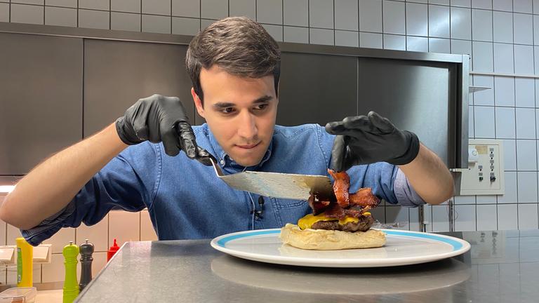 Auf dem Foto sieht man Sherif, wie er sehr konzentriert 2 Speck-Streifen auf den Hamburger legt. Vorher hat er schon ein Burgerpatty mit Käse überbacken und es auch auf die untere Hälfte des Burgerbrötchens gelegt. 
