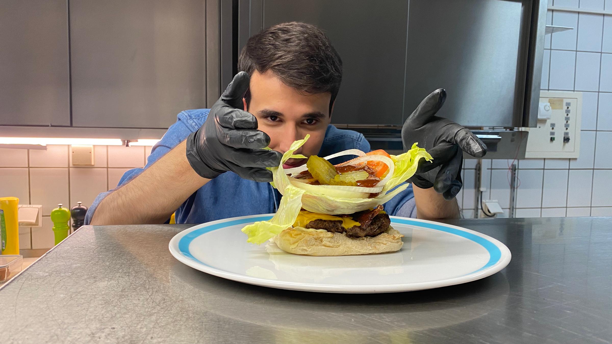 Auf dem Foto sieht man Sherif, wie er ein großes Salatblatt auf dem Hamburger legt. Auf dem Salatblatt liegen außerdem Essiggurkenscheiben, Tomatenscheiben, Zwiebelringe und eine Ladung Ketchup. Sherif trägt schwarze Gummihandschuhe und platziert alles sehr vorsichtig und konzentriert auf dem Burger.