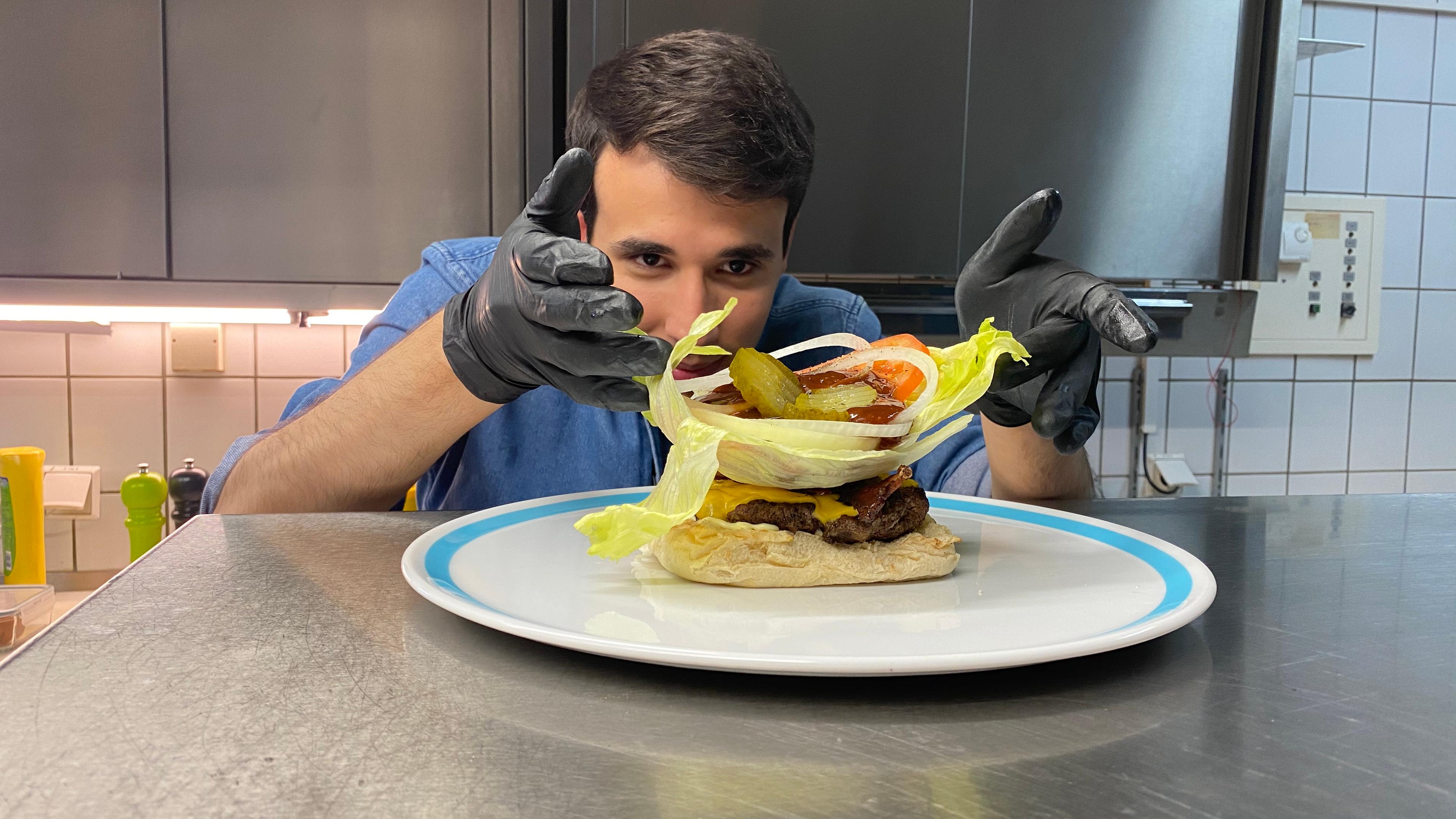 Auf dem Foto sieht man Sherif, wie er ein großes Salatblatt auf dem Hamburger legt. Auf dem Salatblatt liegen außerdem Essiggurkenscheiben, Tomatenscheiben, Zwiebelringe und eine Ladung Ketchup. Sherif trägt schwarze Gummihandschuhe und platziert alles sehr vorsichtig und konzentriert auf dem Burger.