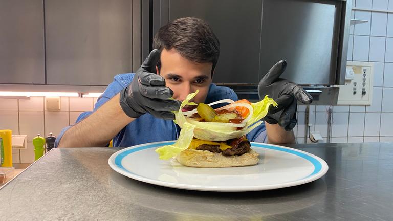 Auf dem Foto sieht man Sherif, wie er ein großes Salatblatt auf dem Hamburger legt. Auf dem Salatblatt liegen außerdem Essiggurkenscheiben, Tomatenscheiben, Zwiebelringe und eine Ladung Ketchup. Sherif trägt schwarze Gummihandschuhe und platziert alles sehr vorsichtig und konzentriert auf dem Burger.