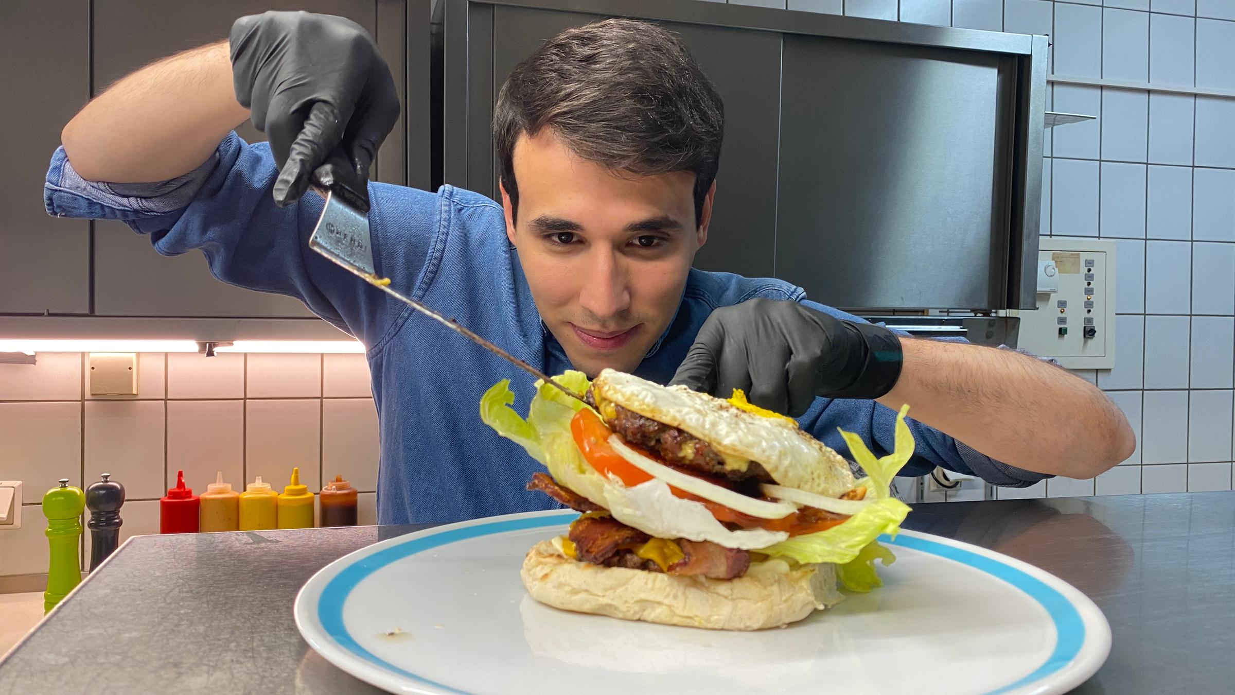 Auf dem Foto platziert Sherif vorsichtig ein zweites Burger-Patty, das mit Käse überbacken wurde. Der Burger ist schon sehr hoch gestapelt, weshalb Sherif zusätzlich alles vorsichtig mit einem Pfannenwender ausbalanciert.