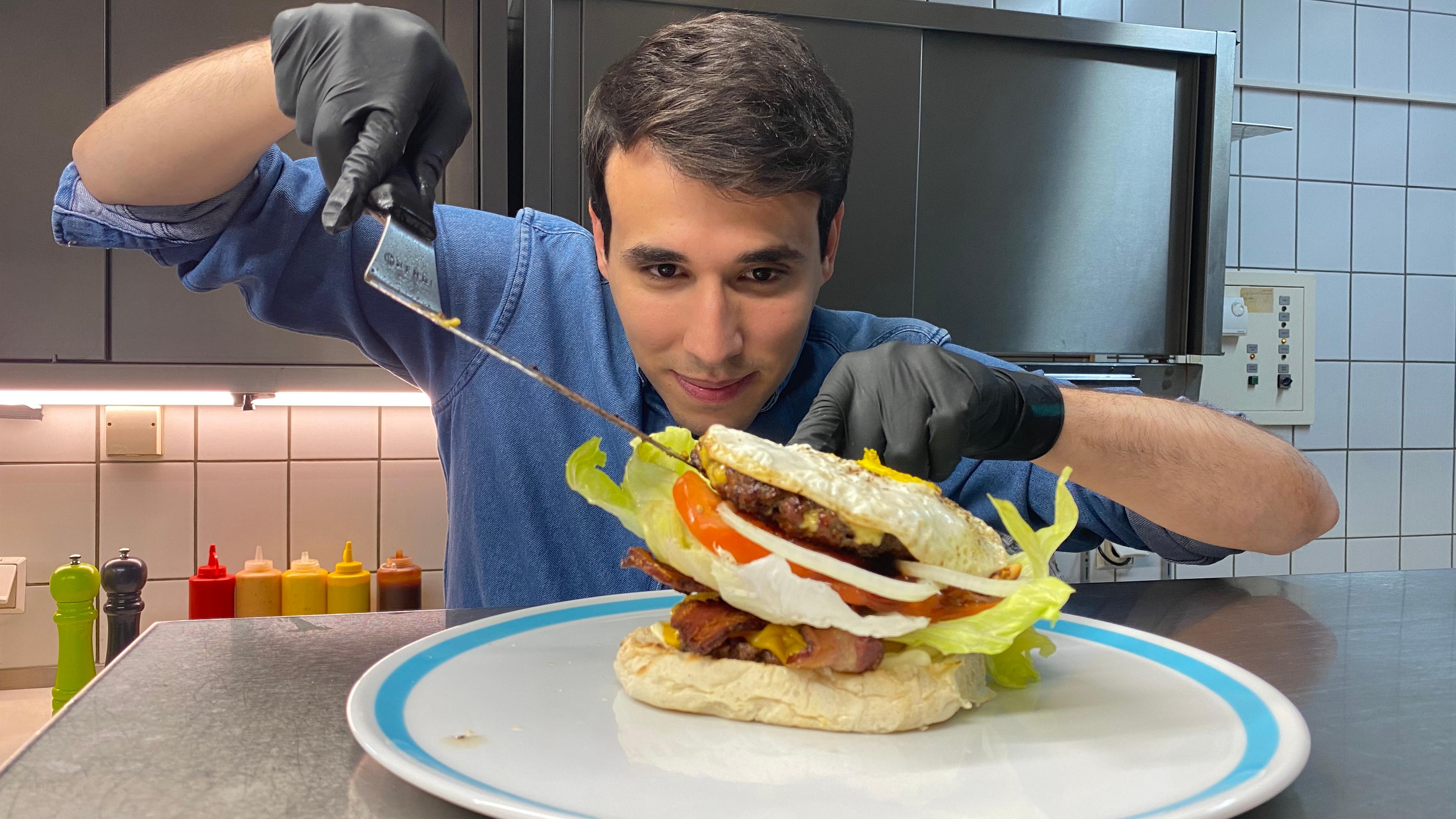 Auf dem Foto platziert Sherif vorsichtig ein zweites Burger-Patty, das mit Käse überbacken wurde. Der Burger ist schon sehr hoch gestapelt, weshalb Sherif zusätzlich alles vorsichtig mit einem Pfannenwender ausbalanciert.