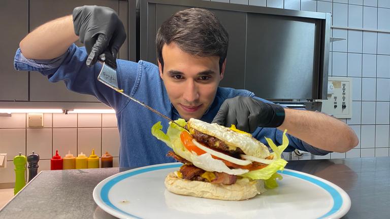 Auf dem Foto platziert Sherif vorsichtig ein zweites Burger-Patty, das mit Käse überbacken wurde. Der Burger ist schon sehr hoch gestapelt, weshalb Sherif zusätzlich alles vorsichtig mit einem Pfannenwender ausbalanciert.