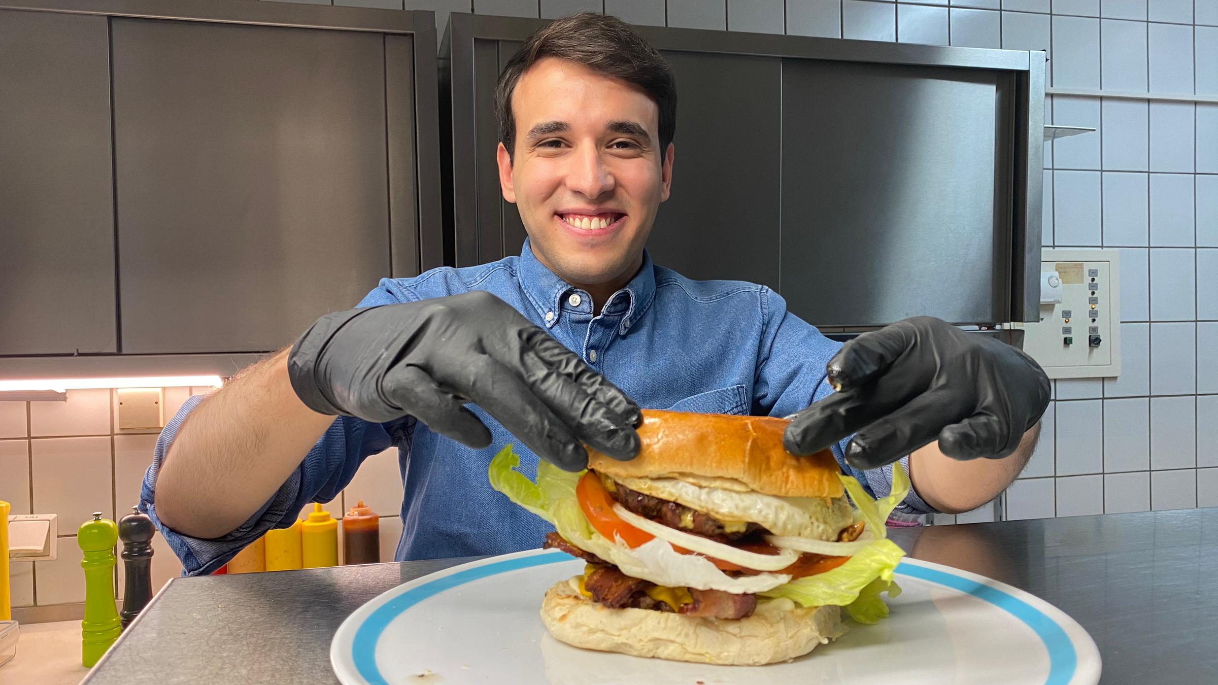 Auf dem Foto legt Sherif mit beiden Händen die obere Hälfte des Burgerbrötchens auf seinen hohen Burger. Dabei grinst er breit.