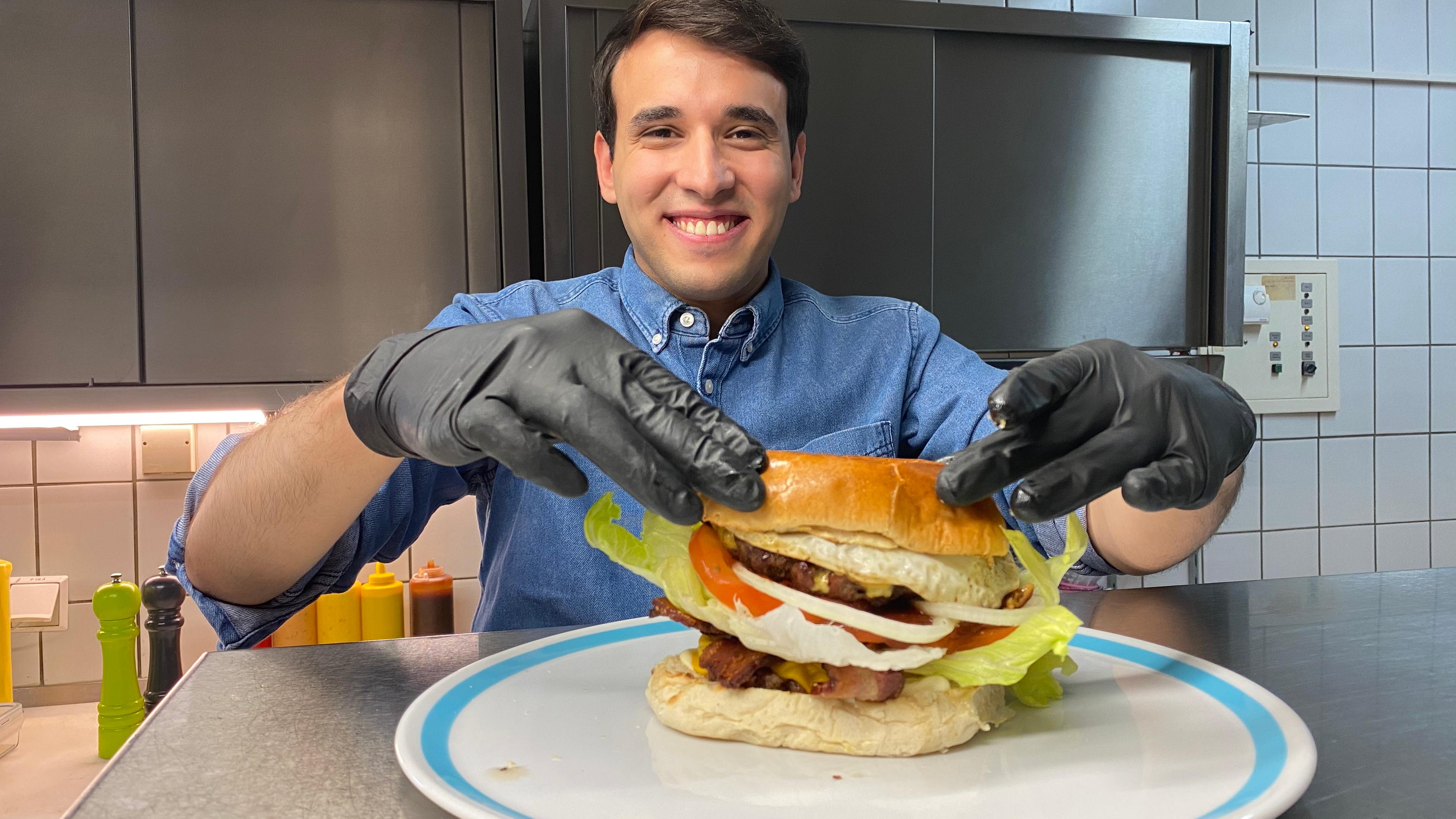 Auf dem Foto legt Sherif mit beiden Händen die obere Hälfte des Burgerbrötchens auf seinen hohen Burger. Dabei grinst er breit.