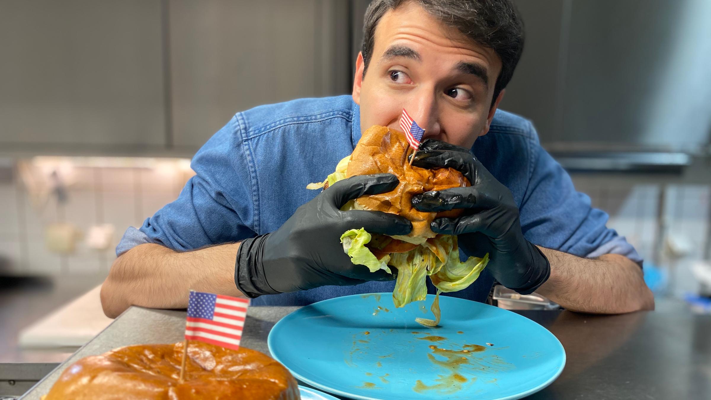 Auf dem Foto probiert Sherif seinen selbstgemachten, sehr großen, amerikanischen Hamburger. Er hält ihn mit beiden Händen fest umschlossen und beißt rein. Er schaut zur Seite und hat seine Augenbrauen hochgezogen.