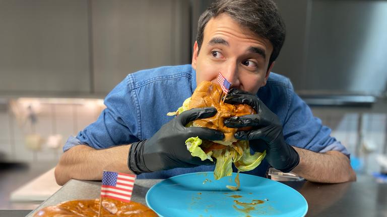 Auf dem Foto probiert Sherif seinen selbstgemachten, sehr großen, amerikanischen Hamburger. Er hält ihn mit beiden Händen fest umschlossen und beißt rein. Er schaut zur Seite und hat seine Augenbrauen hochgezogen.