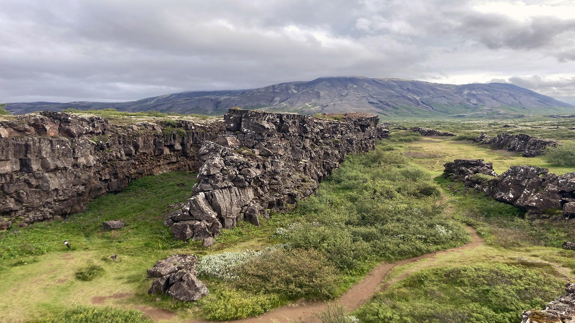 Landschaftsaufnahme vom Grabenbruch von Thingvellir. Inmitten von saftigen, grünen Wiesen türmen sich zwei Felsen auf und bilden einen Graben.