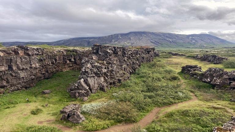 Landschaftsaufnahme vom Grabenbruch von Thingvellir. Inmitten von saftigen, grünen Wiesen türmen sich zwei Felsen auf und bilden einen Graben.