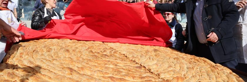 Ein Mann mit schwarzer Kochmütze schlägt ein rotes Tuch auf, das über einer riesigen Teig-Pita liegt. 