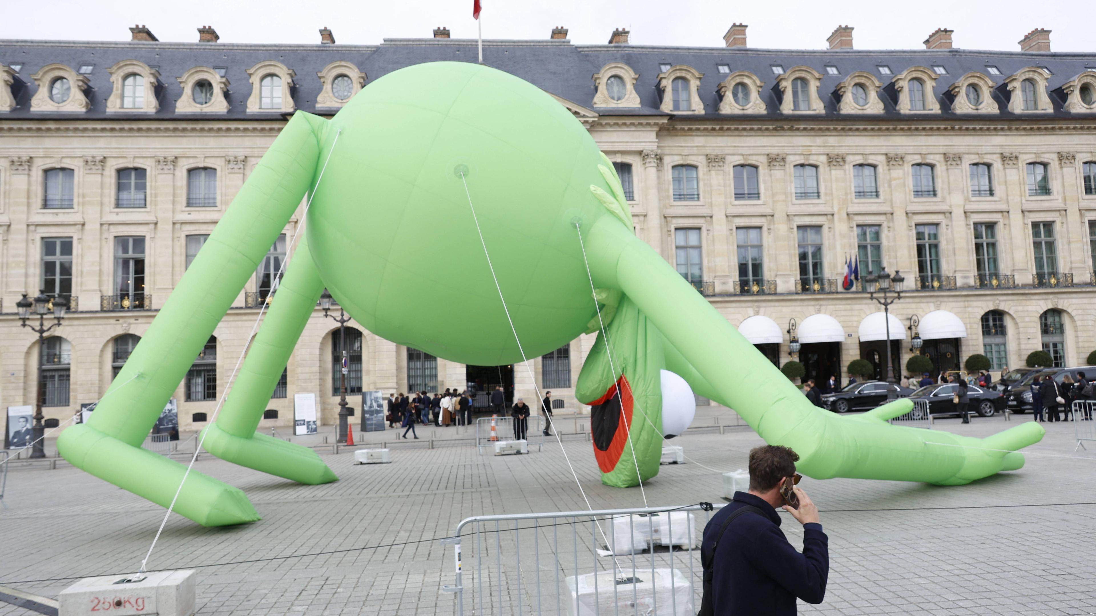 Auf dem Bild ist eine große aufblasbare Figur von dem Frosch Kermit zu sehen. Er ist eingesackt. Sein Kopf berührt deswegen den Boden. Im Hintergrund ist ein Platz in Paris zu erkennen.