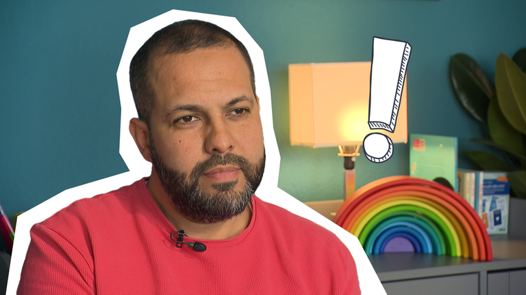 Ein Mann sitzt, hinter ihm steht eine Pflanze und ein Regal, auf dem ein Holz-Regenbogen steht