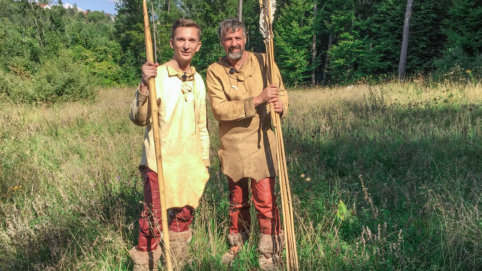 Moderator Eric Mayer steht links im Bild. Er trägt steinzeitiche Kleidung aus Leder und Lederstiefel. In der rechten Hand hält er einen riesigen Speer aus Holz. Rechts nebem ihm steht ebenso gekleidet und mit riesigen Pfeilen in der Hand ein Steinzeitexperte mit ihm auf einer Wiese.