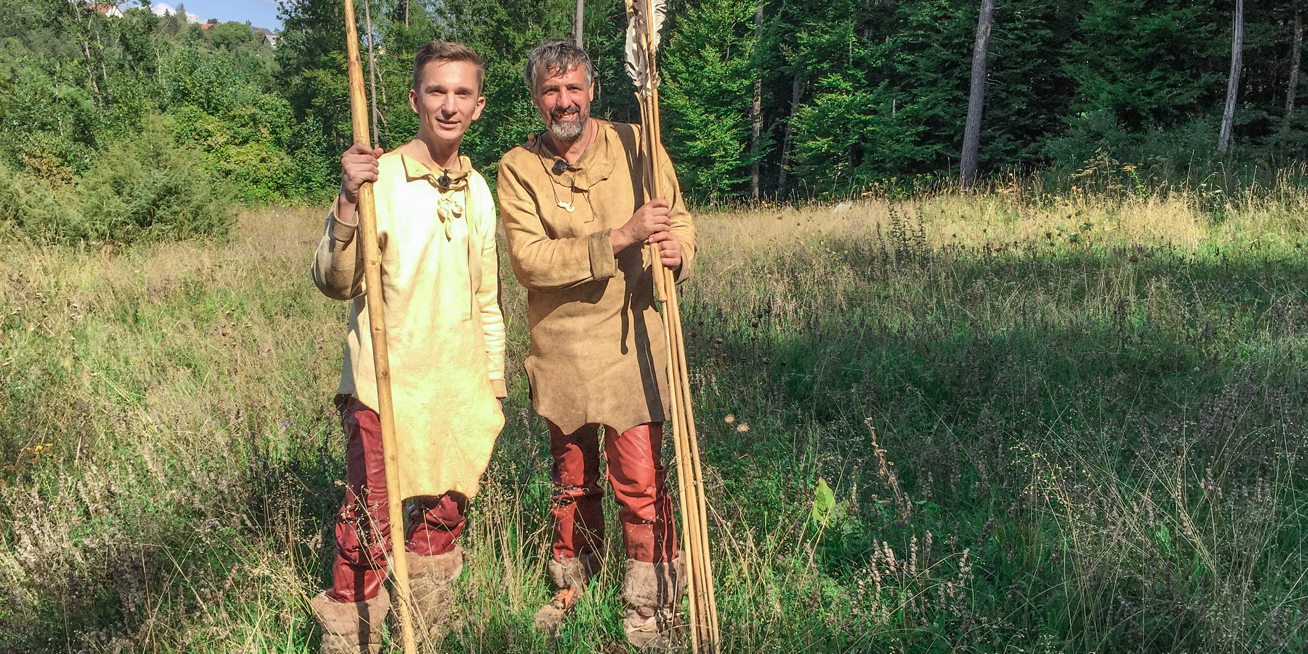 Moderator Eric Mayer steht links im Bild. Er trägt steinzeitiche Kleidung aus Leder und Lederstiefel. In der rechten Hand hält er einen riesigen Speer aus Holz. Rechts nebem ihm steht ebenso gekleidet und mit riesigen Pfeilen in der Hand ein Steinzeitexperte mit ihm auf einer Wiese.