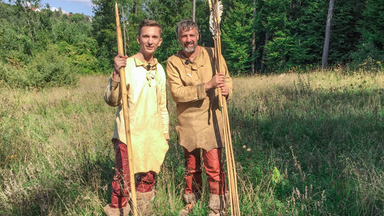 Moderator Eric Mayer steht links im Bild. Er trägt steinzeitiche Kleidung aus Leder und Lederstiefel. In der rechten Hand hält er einen riesigen Speer aus Holz. Rechts nebem ihm steht ebenso gekleidet und mit riesigen Pfeilen in der Hand ein Steinzeitexperte mit ihm auf einer Wiese.
